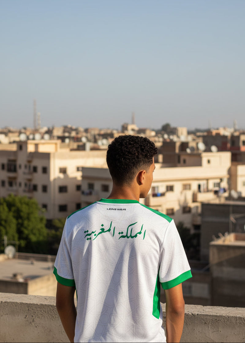 Back view of a Moroccan supporter wearing the Lions Wear Moroccan AURA jersey with Arabic text ‘Kingdom of Morocco’ — rooftop city background.