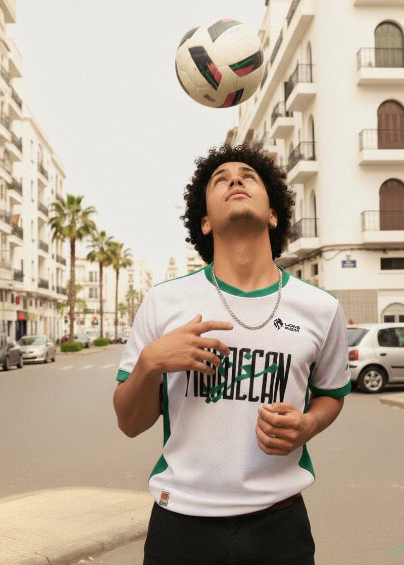 Young Moroccan football fan wearing the Lions Wear Moroccan AURA jersey, balancing a ball on a street with palm trees and white buildings — CAN 2025 supporter style.